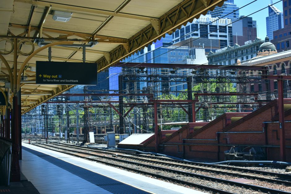 A view of a busy railway station platform with multiple train tracks running parallel to each other, framed by a yellow canopy overhead. The platform surface is paved with concrete, and there is a tac